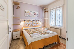 Une chambre à coucher avec un lit, un radiateur, une fenêtre avec des rideaux, un mur orné d’un tableau floral et une étagère avec des vêtements suspendus.