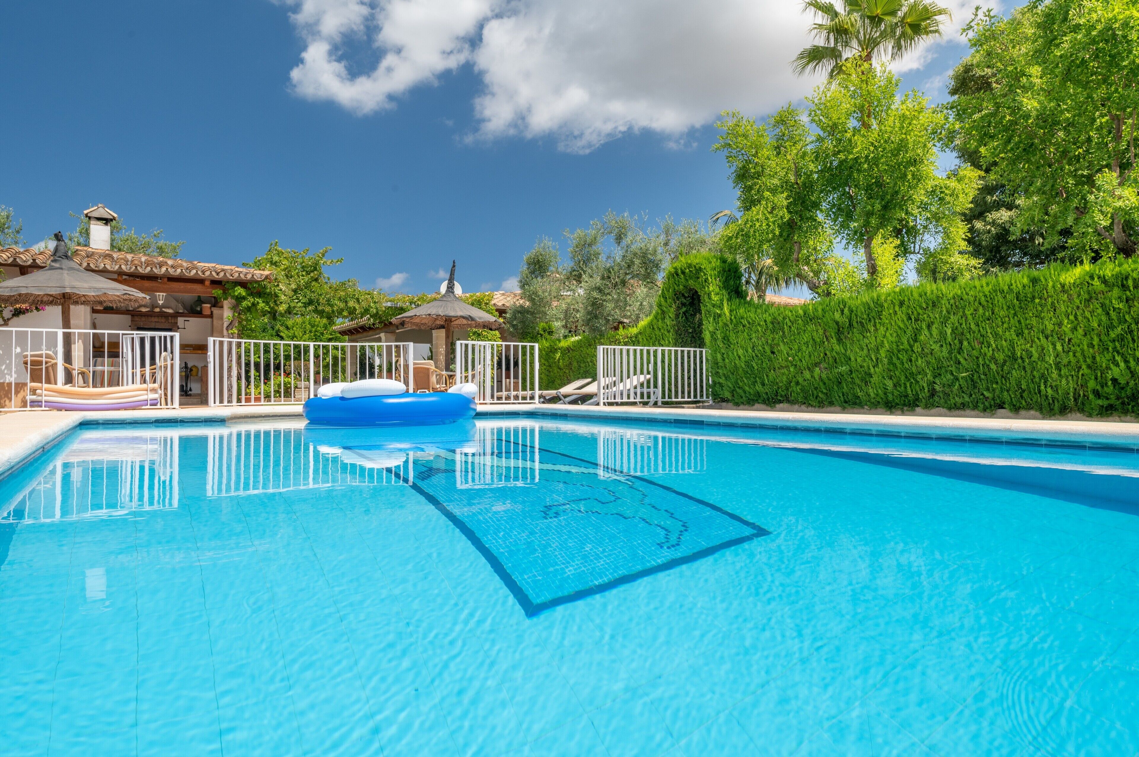 A clear blue pool with a floating ring, surrounded by a white fence, and a building with a tiled roof in the background.