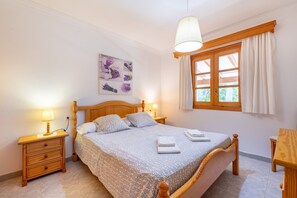 A bedroom with a wooden bed, two bedside tables with lamps, a window with curtains, and a wall-mounted artwork. -  Villa la Rafal with Mountain View, Pool, Wi-Fi, A/C, Terrace & Garden (Pollensa)