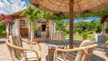 An outdoor patio with wicker chairs, a glass table, and a thatched umbrella.