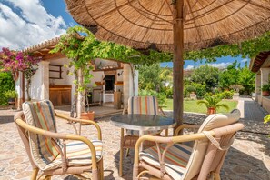 An outdoor patio with wicker chairs, a glass table, and a thatched umbrella. - Villa la Rafal with Mountain View, Pool, Wi-Fi, A/C, Terrace & Garden (Pollensa)