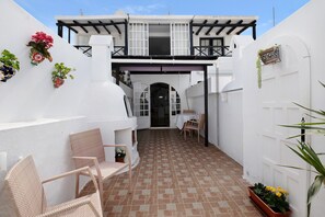 A white building with a balcony, a tiled patio, wicker chairs, and potted plants.