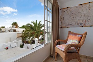 A balcony with a wicker chair, a white wall with a map, and a view of palm trees and buildings.