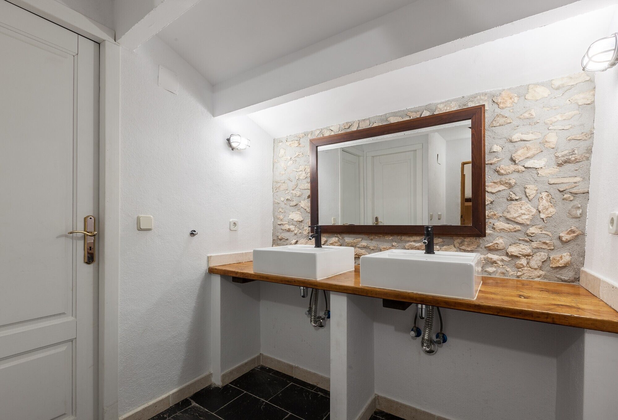 Un baño con dos lavamanos, una encimera de madera, una pared de piedra y una puerta blanca.