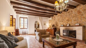 A living room with a fireplace, a chandelier, and a stone wall. - Pet-Friendly Villa Masía Can Trabal with Mountain View, Pool, Jacuzzi, Wi-Fi, A/C, Terrace & Garden (Olèrdola)