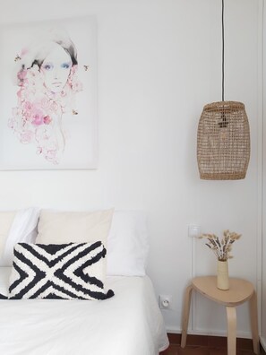A bedroom with a bed, a black and white patterned pillow, a white wall, a hanging woven light fixture, and a small wooden stool with a vase of dried flowers.