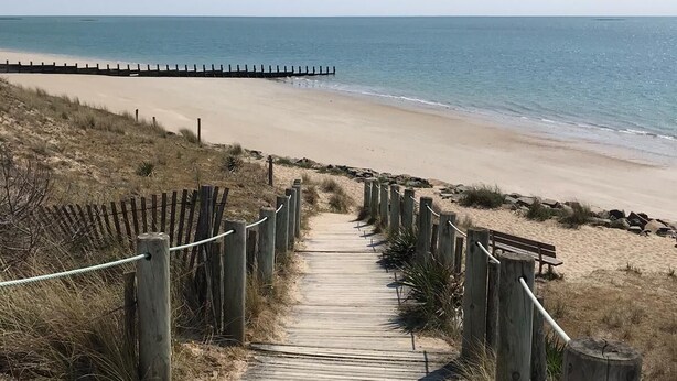 Ein hölzerner Steg führt zu einem Strand, begrenzt auf einer Seite von einem Holzzaun und auf der anderen Seite von einem hölzernen Steg.