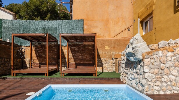 Une piscine avec une terrasse en bois et deux chaises en bois, entourée de murs en pierre et d’un bâtiment à la façade jaune.