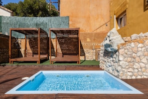 Ein Schwimmbecken mit Holzterrasse und zwei Holzstühlen, umgeben von Steinmauern und einem Gebäude mit gelber Fassade.