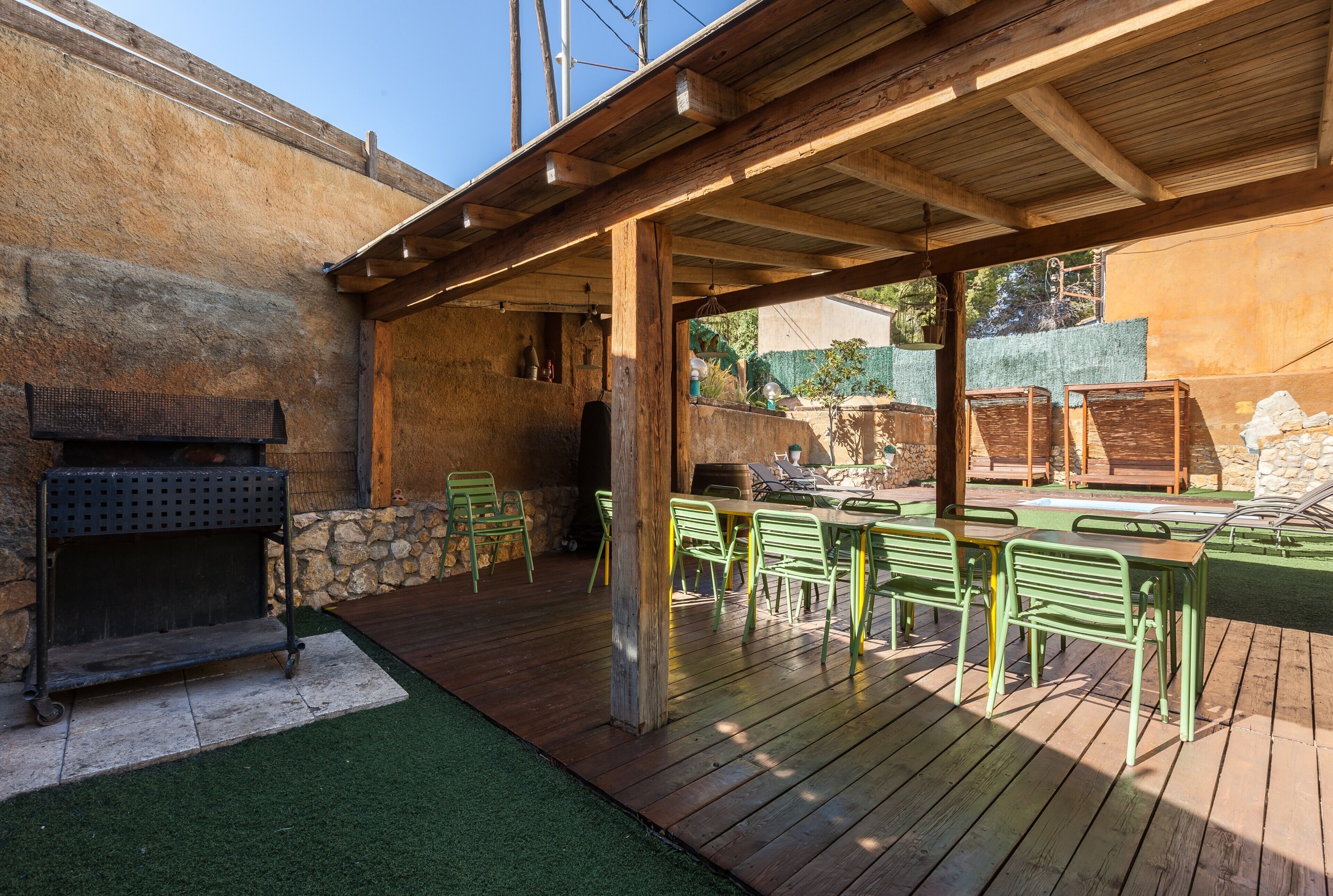 An outdoor patio with a wooden deck, green metal chairs, a grill, and a stone wall.