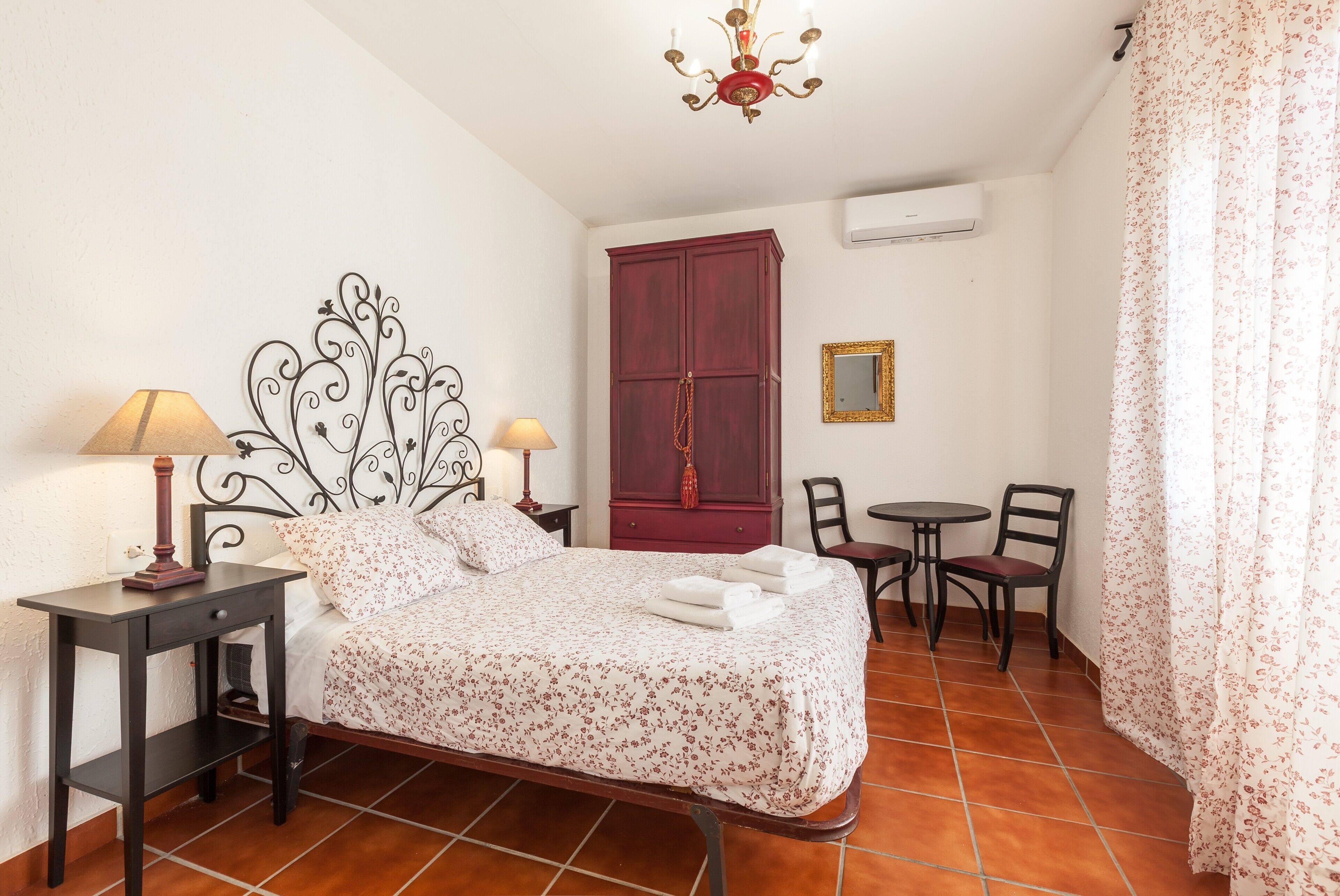 A bedroom with a bed, two bedside tables with lamps, a wardrobe, a small table with chairs, and a ceiling light fixture.