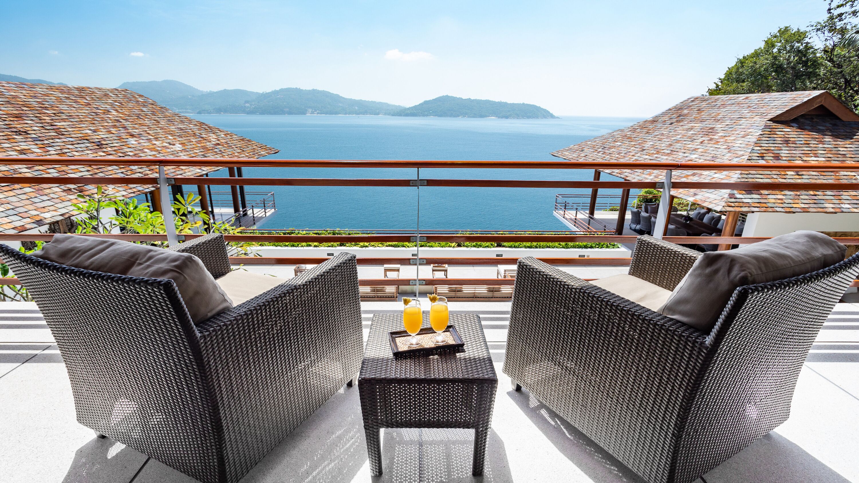 A terrace with wicker furniture and a table, overlooking a body of water and mountains.