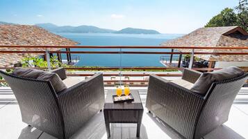 A terrace with wicker furniture and a table, overlooking a body of water and mountains.