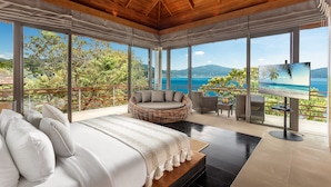 Une chambre avec un grand lit, un canapé en osier, une télévision et un balcon avec vue sur la mer.