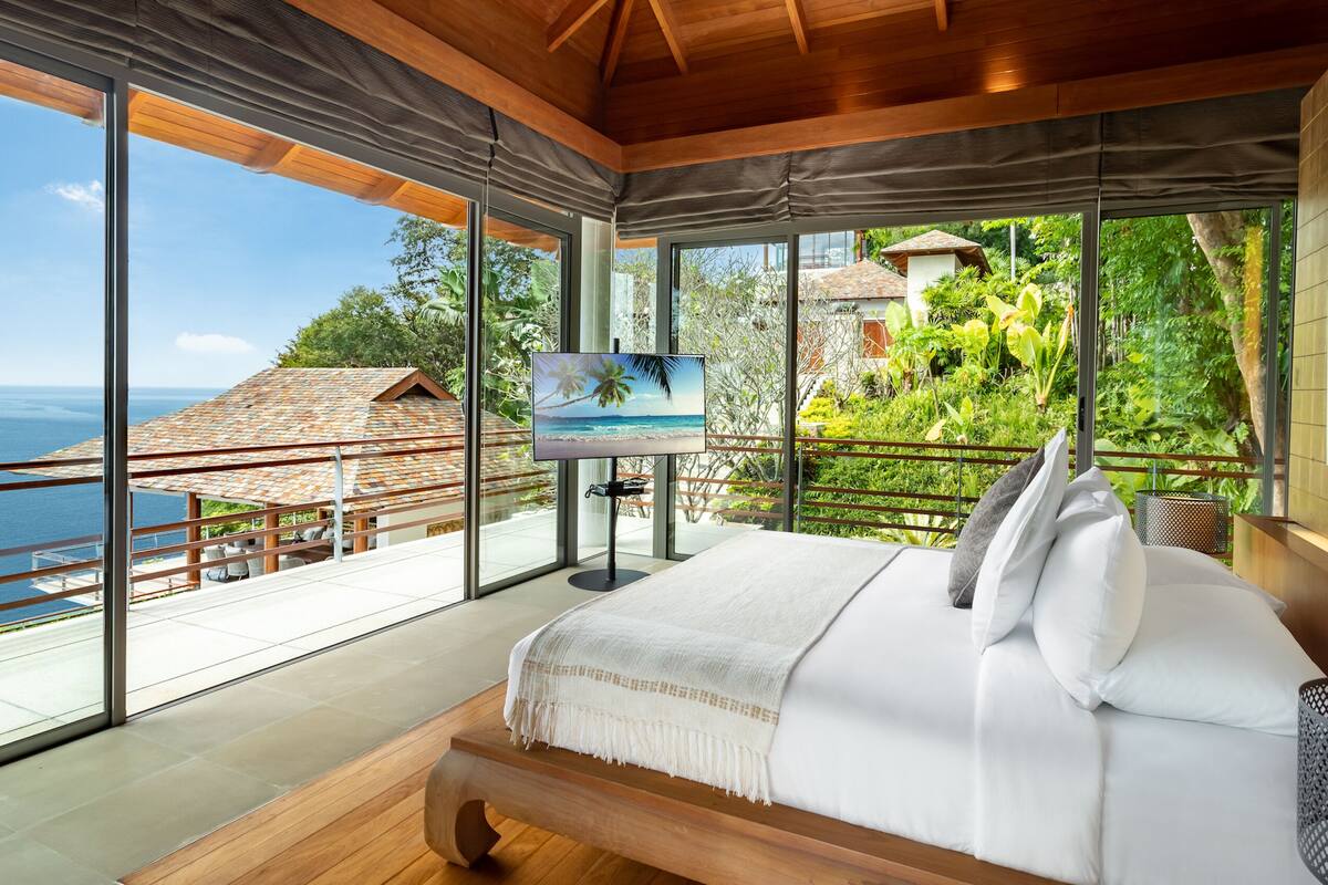Een ruime slaapkamer met een groot bed, een televisie en een balkon met uitzicht op zee en groen.