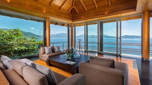 Un intérieur moderne avec un plafond en bois, de grandes fenêtres offrant une vue sur la mer et les collines, un ensemble de canapés et une table basse agrémentée d’objets décoratifs.