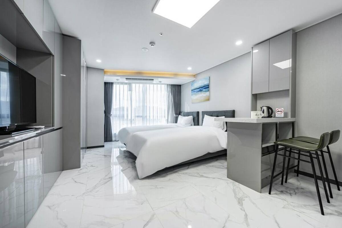 A modern hotel room with a large bed, a desk with a chair, a television, and a view of the city through large windows.