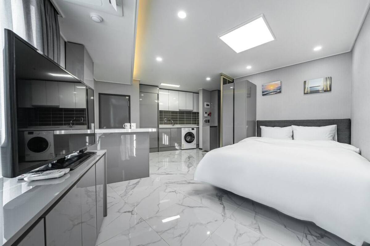 A modern hotel room with a large bed, a separate kitchenette, a built-in washer and dryer, and a bathroom with a shower and toilet.