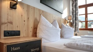 Une chambre d’hôtel avec une tête de lit en bois, du linge de lit blanc, une table de chevet avec une plaque nominative et une fenêtre donnant sur un beau paysage.