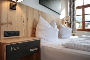 Une chambre d’hôtel avec une tête de lit en bois, du linge de lit blanc, une table de chevet avec une plaque nominative et une fenêtre donnant sur un beau paysage.