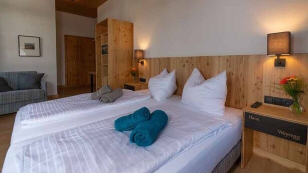A hotel room with a wooden headboard, two beds, a nightstand with a lamp, and a small desk with a vase of flowers.