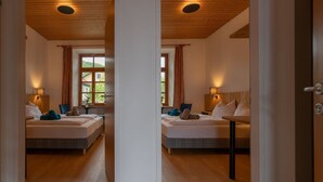 A mirrored view of a hotel room with a wooden ceiling, large windows with curtains, a bed with pillows and a blanket, a bedside table with a lamp, and a wooden floor.