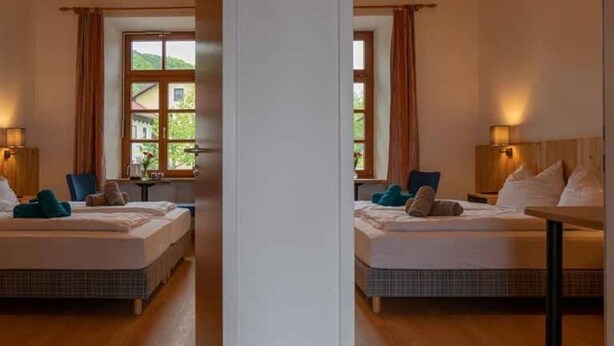 Ein Hotelzimmer mit zwei Betten, Holzboden, einem Fenster mit Vorhängen und einer Lampe auf dem Nachttisch.