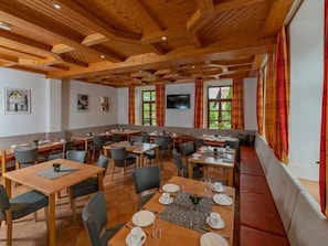 A dining area with wooden tables and chairs, a large window with curtains, and a television on the wall.