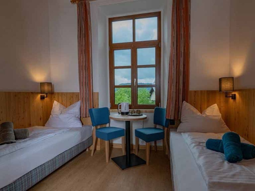 Une chambre d’hôtel avec deux lits, une petite table avec des chaises, une fenêtre avec des rideaux et des lampes fixées au mur.