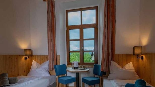 Hotellihuone, jossa on kaksi sänkyä, pieni pöytä tuoleineen, ikkuna verhoineen ja seinään kiinnitetyt lamput.