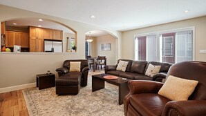 A living room with a brown leather sofa, a coffee table, and a rug. There is a kitchen in the background with wooden cabinets and a refrigerator.