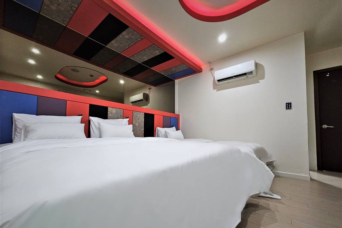 A hotel room with a large bed, a red and blue headboard, an air conditioning unit, and a door with a handle.