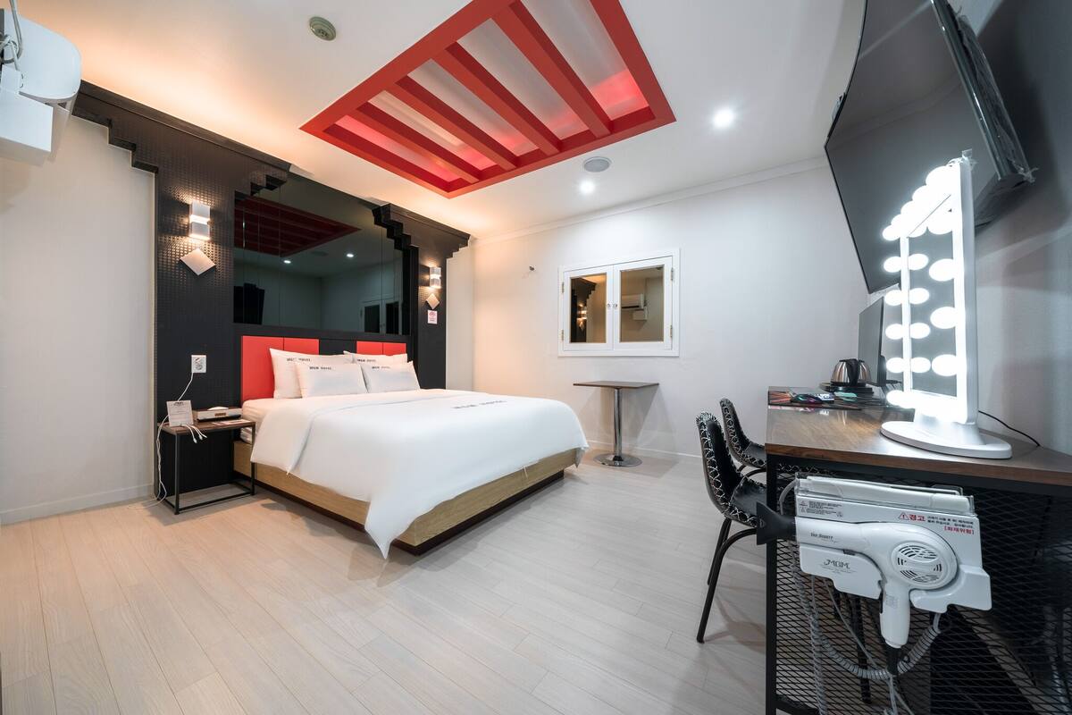 A modern hotel room with a bed, a vanity with lights, a desk, and a chair.