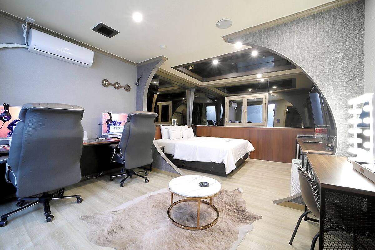 A modern bedroom with a bed, two office chairs, a desk, and a vanity with lights.