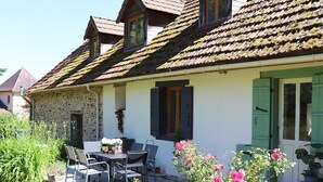 A stone building with a tiled roof, a patio with chairs and a table, and a garden with pink flowers. - La maison d’Etang (Saint-Paul-la-Roche)