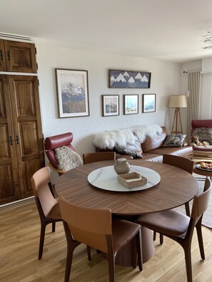 A dining area with a round wooden table, brown leather chairs, and a vase on the table. There is a sofa with a fur throw and pillows in the background.