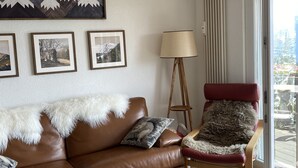A living room with a brown leather sofa, a wooden side table with a fruit bowl, a red chair with a fur throw, a standing lamp, and a view through a glass door.