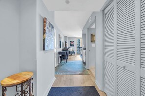 A hallway with a wooden floor, a blue rug, and a wooden stool. There are doors on the right and a dining area with a table and chairs in the background. - Ocean View Style at Kingston Top Location w Pool (Myrtle Beach)