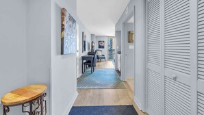 A hallway with a wooden floor, a blue rug, and a wooden stool. There are doors on the right and a dining area with a table and chairs in the background.