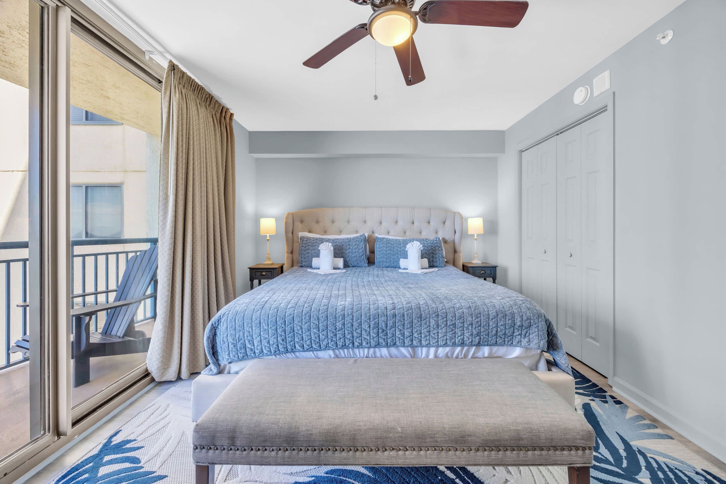 A hotel room with a large bed, a ceiling fan, a balcony with a chair, and a view of the outside.