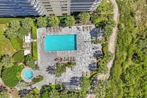 An aerial view of a resort with a central swimming pool, surrounded by lounge areas and palm trees.