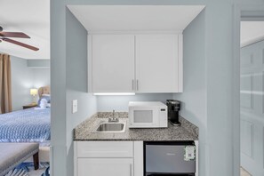 Une chambre d’hôtel compacte comprenant un coin cuisine, un lit avec une couette bleue, un ventilateur de plafond et une fenêtre avec des rideaux.