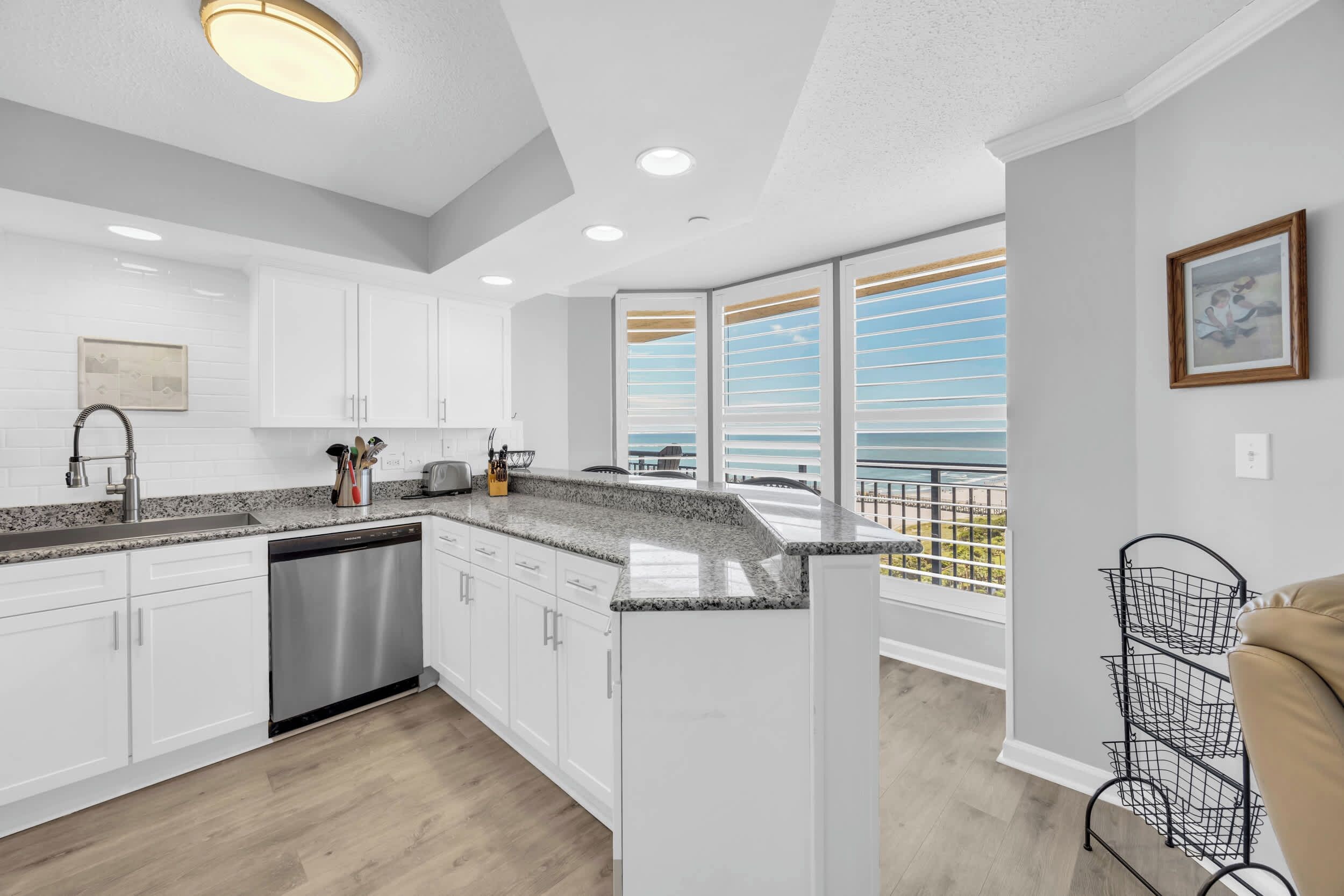 A modern kitchen with white cabinets, granite countertops, and a dishwasher. There is a balcony with a view of the sky and a metal rack on the wall.