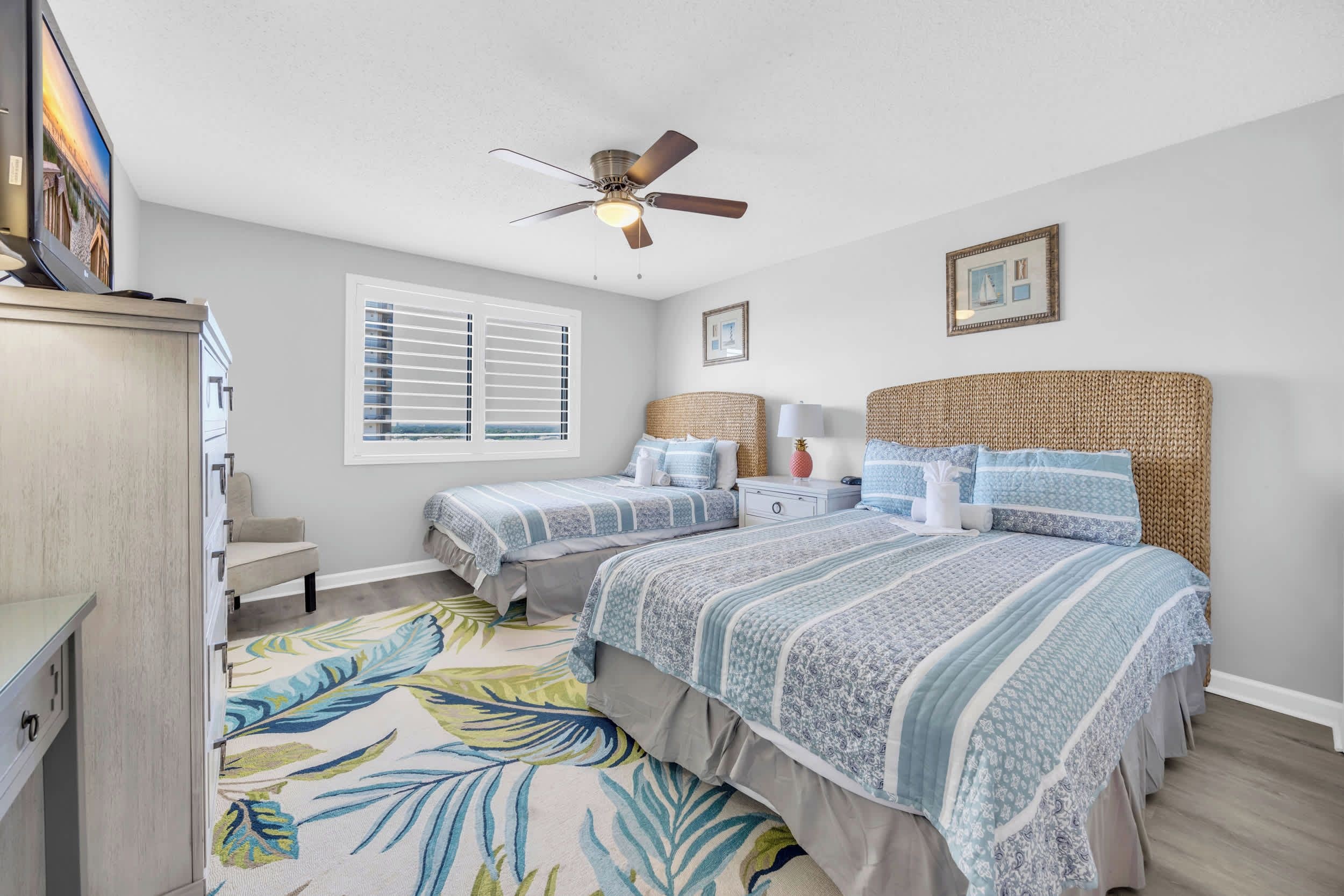 A bedroom with two beds, a ceiling fan, a window with blinds, and a view of the ocean.