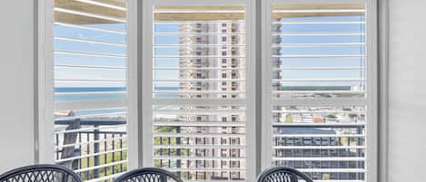 A balcony with white shutters, a view of a high-rise building, and two chairs.