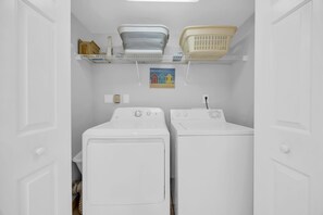 A laundry room with two white washing machines, a shelf with baskets, and a painting of beach houses.