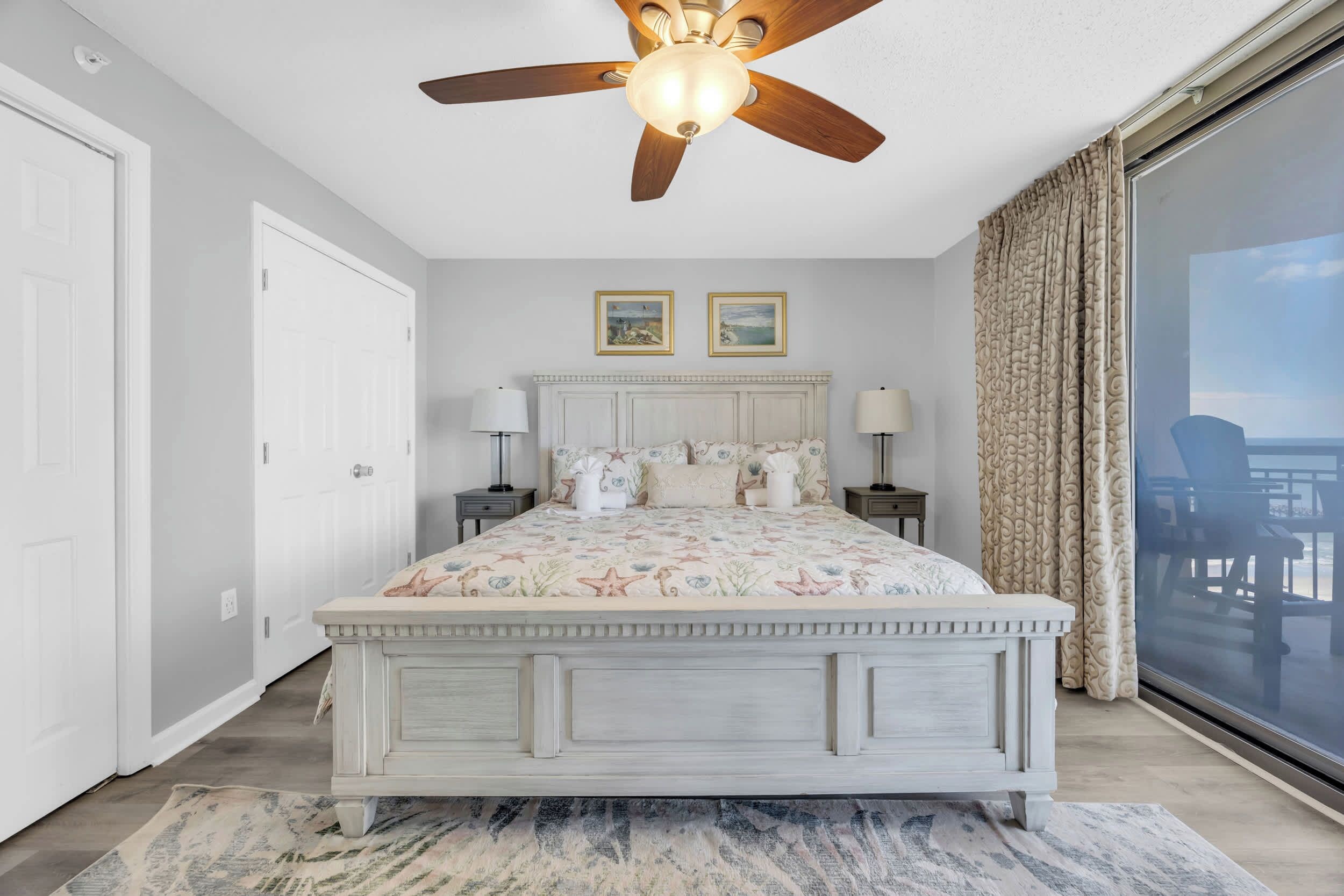 A bedroom with a large bed, a ceiling fan, two wall-mounted lamps, a large window with curtains, and a view of the outdoors.
