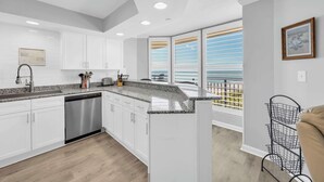 Eine moderne Küche mit weißen Schränken, Granitarbeitsplatten und einem Geschirrspüler. Es gibt einen Balkon mit Blick auf den Himmel und einen Metallregal an der Wand.