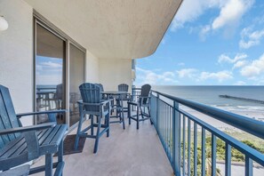 A balcony with chairs and tables overlooking a beach and pier.