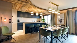 A modern dining area with a wooden ceiling, a kitchen in the background, and a dining table set for six. - Méribel: 140sqm Apartment, 4 Bedrooms, Ski Access, Fireplace, WIFI, Parking (Méribel)